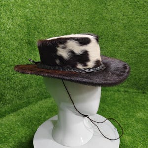 Pode incluir: Um chap&eacute;u de cowboy com aba marrom e copa com estampa de pele de vaca preta e branca. O chap&eacute;u tem uma faixa de couro tran&ccedil;ado e uma tira de queixo. O chap&eacute;u &eacute; exibido em uma cabe&ccedil;a de manequim branca sobre um fundo verde.