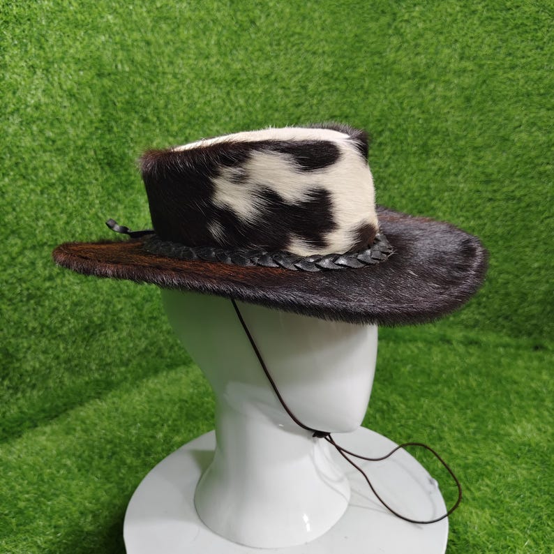 Pode incluir: Um chap&eacute;u de cowboy com aba marrom e copa com estampa de pele de vaca preta e branca. O chap&eacute;u tem uma faixa de couro tran&ccedil;ado e uma tira de queixo. O chap&eacute;u &eacute; exibido em uma cabe&ccedil;a de manequim branca sobre um fundo verde.
