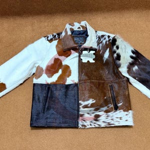 May include: A cowhide jacket with a patchwork design. The jacket features a mix of white, brown, and black cowhide panels. It has a collared neckline, a zipper closure, and two front pockets. The sleeves are long.