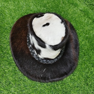 Pode incluir: Um chap&eacute;u redondo com uma copa de pele de vaca e uma aba castanha escura. A copa apresenta um padr&atilde;o a preto e branco, e o chap&eacute;u tem uma faixa de couro tran&ccedil;ado preto. O chap&eacute;u est&aacute; sobre um fundo verde.