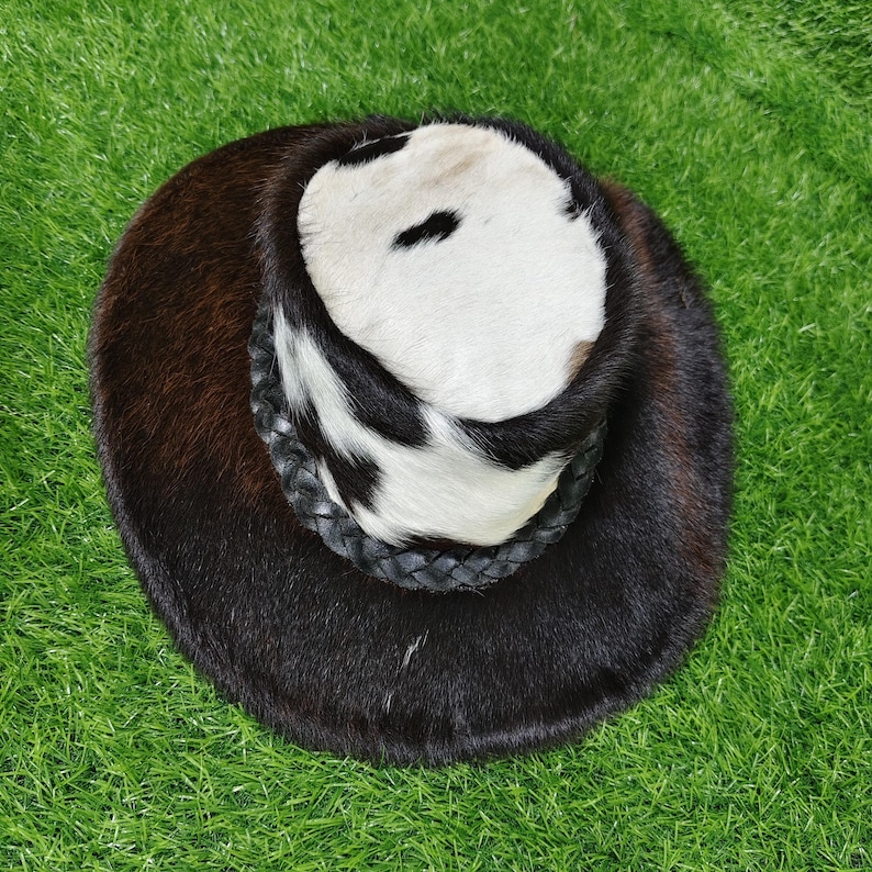 Pode incluir: Um chap&eacute;u redondo com uma copa de pele de vaca e uma aba castanha escura. A copa apresenta um padr&atilde;o a preto e branco, e o chap&eacute;u tem uma faixa de couro tran&ccedil;ado preto. O chap&eacute;u est&aacute; sobre um fundo verde.
