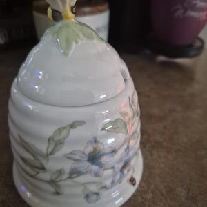 May include: White ceramic honey pot with a bee-shaped lid handle. The pot features a floral design with blue and green accents. The pot has a beehive shape and is designed for serving honey.