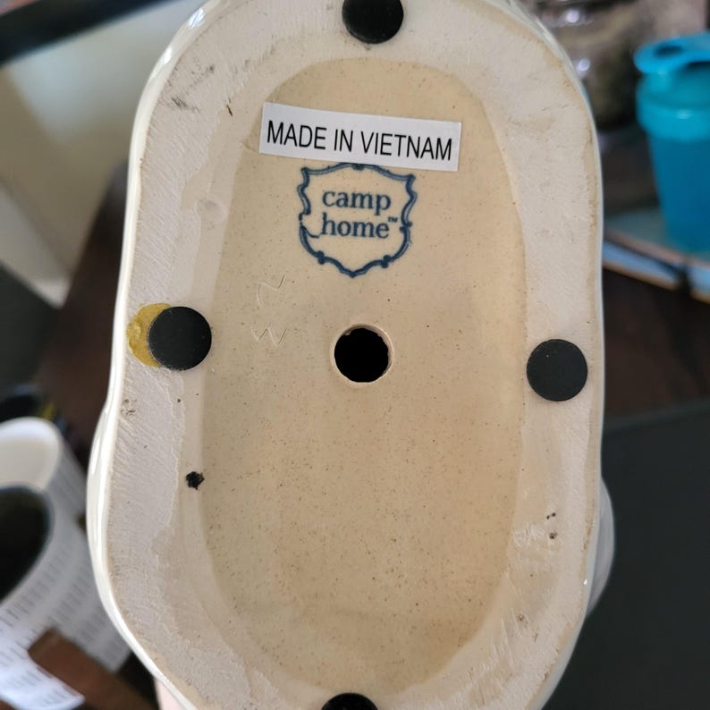 May include: The bottom of a white ceramic figurine with a blue and white logo that reads "camp home" and the text "Made in Vietnam".