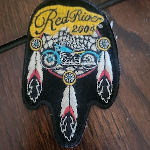 May include: Black embroidered patch with the text "Red River 2004". The patch features a motorcycle design in blue and white, surrounded by decorative elements including feathers and circular patterns.