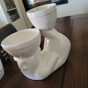 May include: White ceramic planter shaped like a cat with two white ceramic bowls on top. The bowls have black speckles.