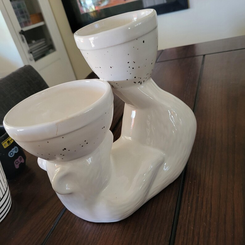 May include: White ceramic planter shaped like a cat with two white ceramic bowls on top. The bowls have black speckles.
