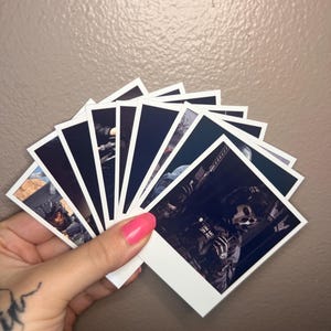 May include: A collection of square, white-bordered photographs featuring images of a character in a military uniform and skull mask. The photos are held in a hand with pink nail polish against a neutral background.