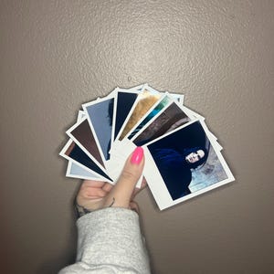 May include: A collection of Polaroid photos featuring a person with dark hair and a black shirt. The photos are arranged in a fan shape and held in a hand with pink nail polish.