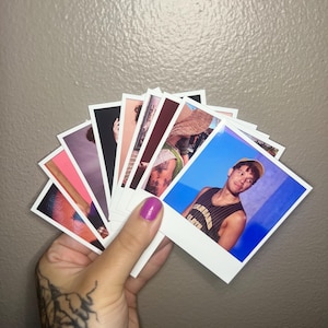 May include: A hand holding a fan of polaroid-style photographs. The photos feature various images, including portraits of people. The hand has a tattoo on the wrist and is painted with purple nail polish. The background is a solid, neutral color.