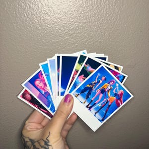 May include: A collection of square, white-bordered photo prints featuring colorful images of people in various poses. The prints are held in a hand with a purple painted nail. The background is a solid, neutral color.