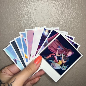 May include: A hand holding a fan of square-shaped art prints. The prints feature colorful illustrations of figures in various poses. The prints have white borders and are held against a neutral background.