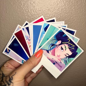 May include: A collection of square art prints featuring colorful illustrations. The prints showcase a variety of designs, including portraits and abstract art. The prints are held in a hand with an orange painted nail.
