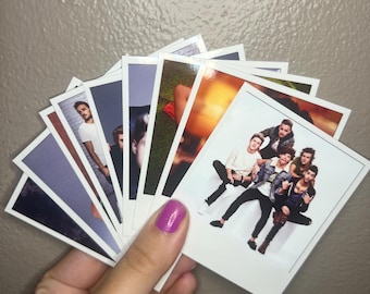 Op de afbeelding: Een hand die een waaier van foto's in polaroidstijl vasthoudt. De foto's bevatten afbeeldingen van een muziekgroep. De foto's hebben witte randen en zijn uitgewaaierd om de afbeeldingen weer te geven. De achtergrond is neutraal van kleur.