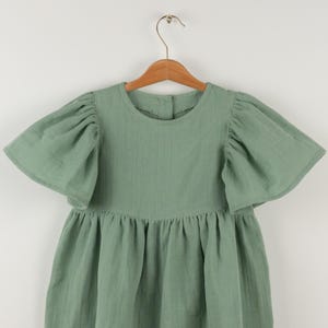 May include: A sage green dress with short, puffed sleeves and a gathered skirt, hanging on a wooden hanger. The dress has a round neckline and a button closure at the back. The fabric appears to be a lightweight cotton.