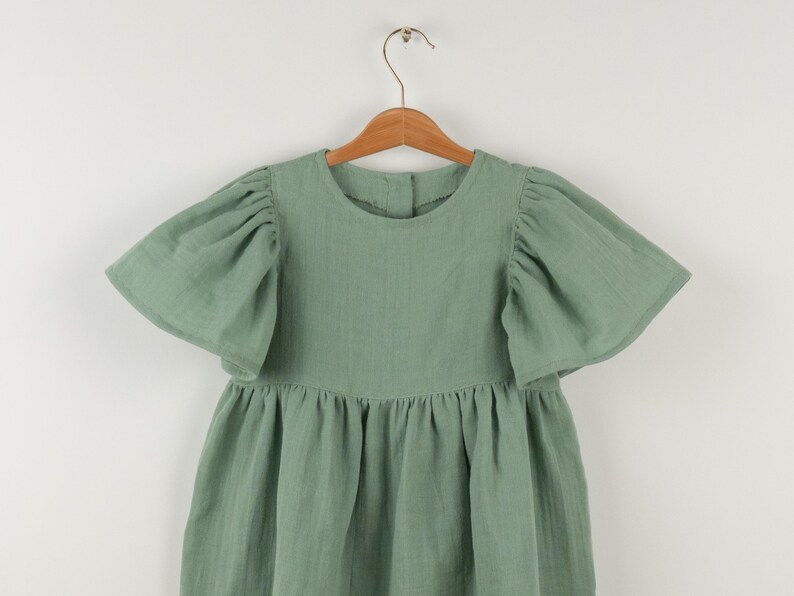 May include: A sage green dress with short, puffed sleeves and a gathered skirt, hanging on a wooden hanger. The dress has a round neckline and a button closure at the back. The fabric appears to be a lightweight cotton.