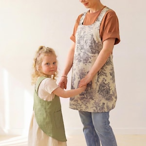 May include: A beige apron featuring a dark gray toile de Jouy pattern with pastoral scenes and a sage green apron with a square pocket. Both aprons have a cross-back design, wide shoulder straps, and a sleeveless silhouette that covers the torso.