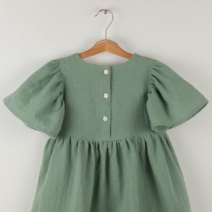May include: Sage green dress with short, puffed sleeves and a gathered skirt. The dress features a button-down back and is hanging on a wooden hanger. The fabric has a textured appearance.