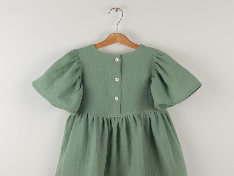 May include: Sage green dress with short, puffed sleeves and a gathered skirt. The dress features a button-down back and is hanging on a wooden hanger. The fabric has a textured appearance.