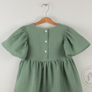 May include: Sage green dress with short, puffed sleeves and a gathered skirt, hanging on a wooden hanger. The dress has a button-up back and a round neckline. The dress is made of a textured fabric. The Ela Patterns logo is visible.