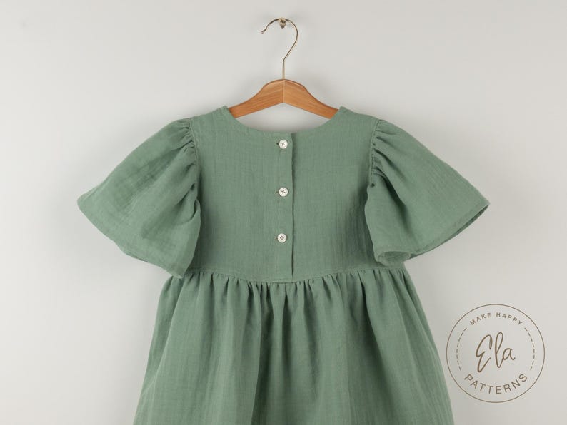 May include: Sage green dress with short, puffed sleeves and a gathered skirt, hanging on a wooden hanger. The dress has a button-up back and a round neckline. The dress is made of a textured fabric. The Ela Patterns logo is visible.