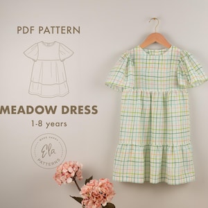 May include: A light green and white plaid Meadow Dress for ages 1-8 years hangs on a wooden hanger. The dress has short, flutter sleeves and a tiered skirt. The image also includes a line drawing of the dress and the text "PDF Pattern".
