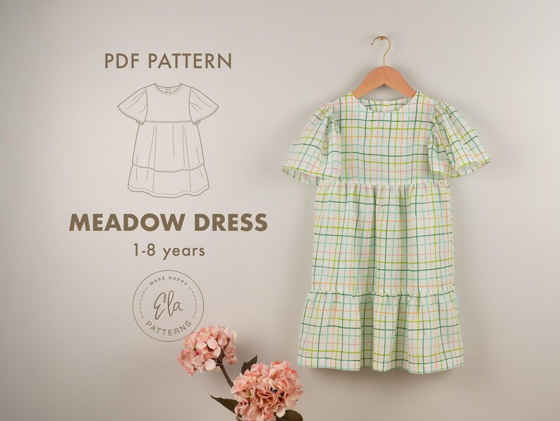 May include: A light green and white plaid Meadow Dress for ages 1-8 years hangs on a wooden hanger. The dress has short, flutter sleeves and a tiered skirt. The image also includes a line drawing of the dress and the text "PDF Pattern".