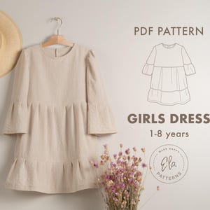 Op de afbeelding: Een beige linnen jurk met een ronde hals en lange mouwen. De jurk heeft een gerimpelde zoom en hangt aan een houten hanger. De jurk is voor meisjes van 1 tot 8 jaar oud. De afbeelding bevat ook een PDF-patroon voor de jurk.
