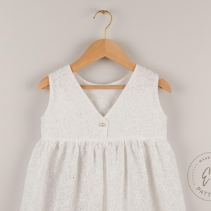 May include: A white sleeveless dress with a V-neck and floral eyelet pattern, hanging on a wooden hanger. The dress has a button closure and a gathered skirt. The dress is from Ela Patterns.