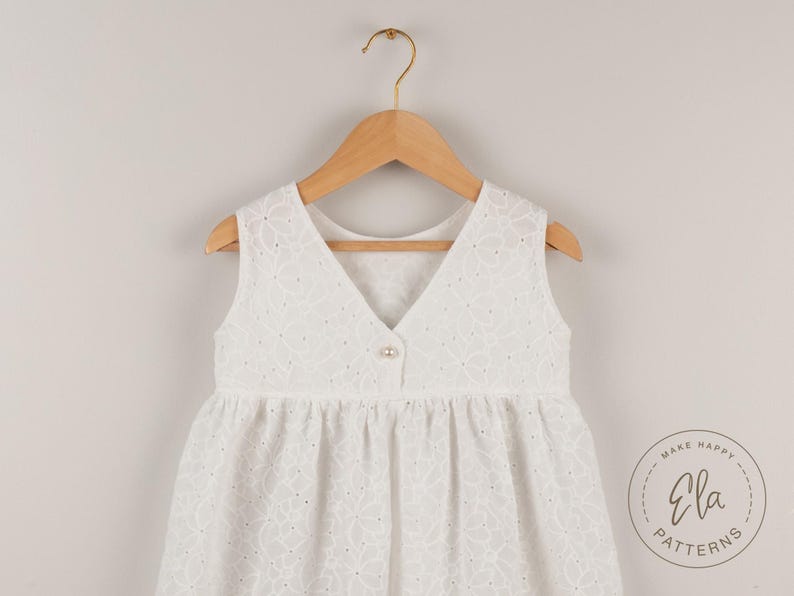 May include: A white sleeveless dress with a V-neck and floral eyelet pattern, hanging on a wooden hanger. The dress has a button closure and a gathered skirt. The dress is from Ela Patterns.