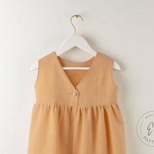 May include: A sleeveless, light brown dress with a V-neck and a single button closure. The dress is hanging on a white hanger against a white background. The dress has a gathered waist and a flared skirt. The logo "Ela Patterns" is visible.