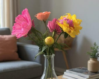 May include: A clear glass vase holds a vibrant bouquet of colorful paper flowers. The arrangement includes large, bright pink, orange, and yellow blooms. The vase sits on a light wood table, with a gray sofa and books in the background.