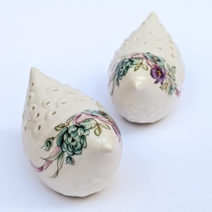 May include: Two off-white ceramic bird-shaped candle holders. Each has a floral design with green, purple, and pink accents. The candle holders have small holes for light to shine through, and a pointed beak.