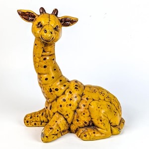 May include: A ceramic giraffe figurine in a seated position. The giraffe is primarily yellow with brown spots and accents. The ears and horns are brown. The figurine has a smiling expression and is set against a white background.
