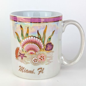 May include: White ceramic mug with a pearlescent finish, featuring a seashell and coral design in pink, gold, and green. The mug has a pink and gold rim and the words "Miami, Fl" in gold script.