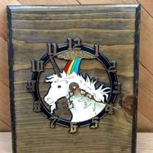 May include: A wooden clock featuring a white unicorn with a rainbow and gold glitter accents. The clock face is black with gold-colored numerals. The wooden frame has a dark brown stain.