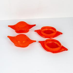 May include: A set of four bright orange glass dishes, each featuring a central circular bowl section flanked by two wing-like extensions with scalloped edges. The translucent glass items are arranged in a two-by-two formation on a white surface.