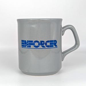 May include: A gray ceramic mug with a curved handle. The word "ENFORCER" is printed in blue block letters across the front. The mug has a simple, classic design and is suitable for hot or cold beverages.