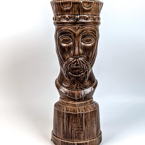 May include: A brown ceramic sculpture of a king's head, featuring a detailed face with a beard and a crown. The sculpture has a textured, wood-like appearance and is mounted on a cylindrical base. The piece is a decorative item.