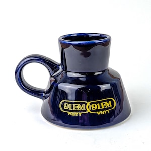May include: A navy blue ceramic mug with a unique shape and a rounded handle. The mug features the text "91FM WHYY" in yellow lettering. The mug has a glossy finish and is designed for beverages.