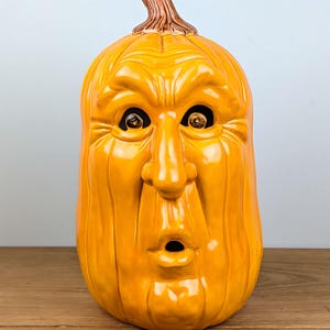 May include: A ceramic pumpkin with a surprised face. The pumpkin is orange with a brown stem and has large eyes and an open mouth. The pumpkin is a decorative item, likely for Halloween.