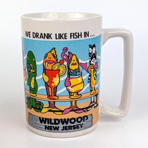 May include: White ceramic mug with a rectangular handle. The mug features a colorful illustration of fish drinking cocktails with the text "WE DRANK LIKE FISH IN... WILDWOOD NEW JERSEY". The background has a blue sky and beach scene.