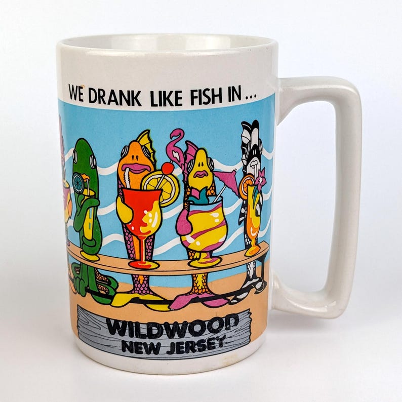 May include: White ceramic mug with a rectangular handle. The mug features a colorful illustration of fish drinking cocktails with the text "WE DRANK LIKE FISH IN... WILDWOOD NEW JERSEY". The background has a blue sky and beach scene.