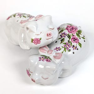 May include: White ceramic pig figurines with pink floral and bow accents. The pigs are in a sleeping position, with closed eyes and delicate painted details. A charming decorative set for a kitchen or dining area.