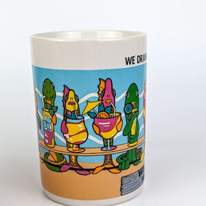 May include: White ceramic mug with a colorful cartoon illustration of fish-like creatures at a bar, each holding a cocktail. The mug has a blue background with the words "WE DRAW" on the side.
