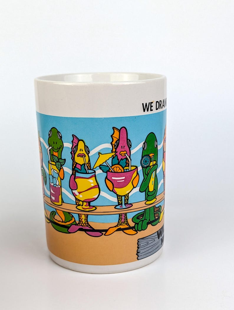 May include: White ceramic mug with a colorful cartoon illustration of fish-like creatures at a bar, each holding a cocktail. The mug has a blue background with the words "WE DRAW" on the side.