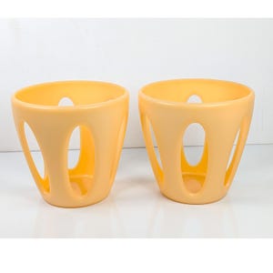 May include: Two yellow-orange, open-sided, cylindrical containers. Each container has a rounded top and base, with several oval-shaped cutouts along the sides. The containers are made of a smooth, solid material and sit on a white surface.
