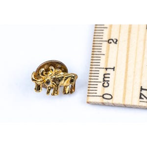 May include: A small, gold-colored bull pin. The pin is approximately 0.75 inches long and features a detailed design of a bull. The pin has a standard pin back for attachment. The bull is standing on all fours.