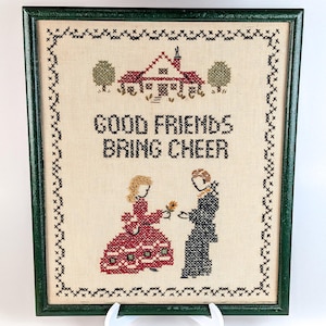 May include: Cross-stitch artwork in a green frame. The piece features a house, trees, and two figures, with the text "GOOD FRIENDS BRING CHEER." The figures are exchanging a flower. The frame is supported by a white easel.