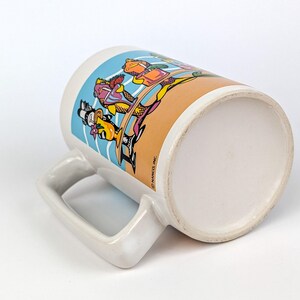 May include: White ceramic mug with a cartoon design featuring characters in colorful outfits. The mug has a large handle and a wide base. The design includes blue, orange, yellow, and purple colors.