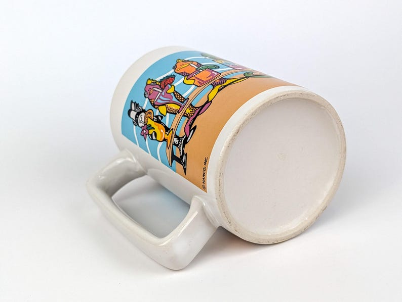 May include: White ceramic mug with a cartoon design featuring characters in colorful outfits. The mug has a large handle and a wide base. The design includes blue, orange, yellow, and purple colors.